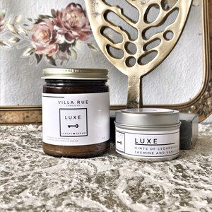 May include: Two candles in glass jars with silver lids. The larger jar is labelled "Villa Rue Candle Co. Luxe Serene Dream" and the smaller jar is labelled "Luxe Hints of Cedarwood Jasmine and Vanilla".