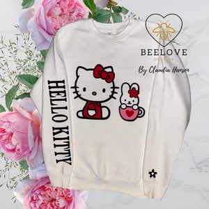 Pode incluir: Camisola branca com Hello Kitty e um coelho com laços e detalhes vermelhos. A manga esquerda tem a inscrição "HELLO KITTY" em preto. Uma pequena flor preta está na outra manga. O design inclui o texto "BEE LOVE DESIGNS By Claudia Hansen".