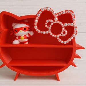 K&ouml;nnte beinhalten: Ein rotes Regal in Form von Hello Kittys Kopf, mit zwei Regalb&ouml;den und einer mit Perlen besetzten Schleife. Eine Hello Kitty-Figur im Superhelden-Kost&uuml;m befindet sich auf dem oberen Regal. Das Regal hat einen dreibeinigen Sockel.