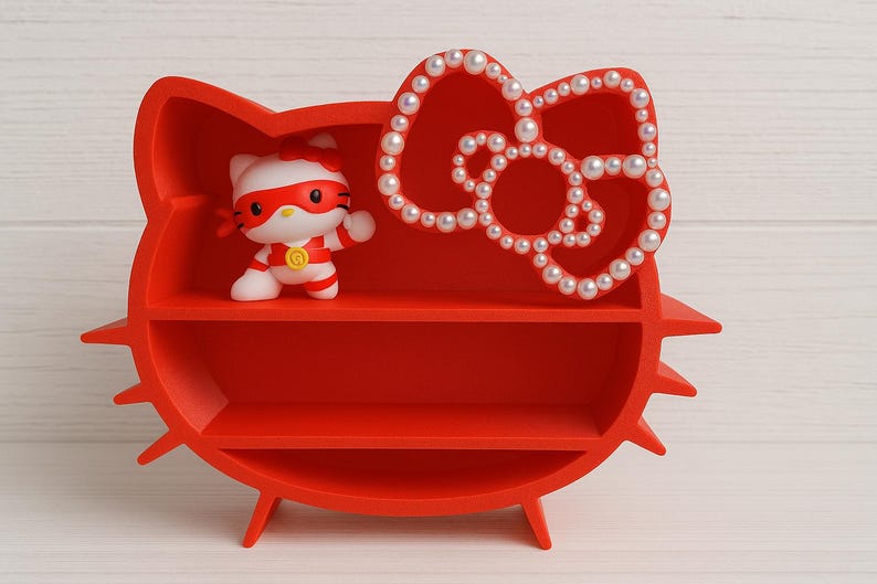 K&ouml;nnte beinhalten: Ein rotes Regal in Form von Hello Kittys Kopf, mit zwei Regalb&ouml;den und einer mit Perlen besetzten Schleife. Eine Hello Kitty-Figur im Superhelden-Kost&uuml;m befindet sich auf dem oberen Regal. Das Regal hat einen dreibeinigen Sockel.
