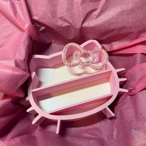K&ouml;nnte beinhalten: Ein rosa Organizer mit drei F&auml;chern in Form eines Zeichentrickkatzenkopfes. Der Organizer hat eine schleifenf&ouml;rmige Verzierung mit kleinen, irisierenden Perlen. Er liegt auf rosa Seidenpapier.