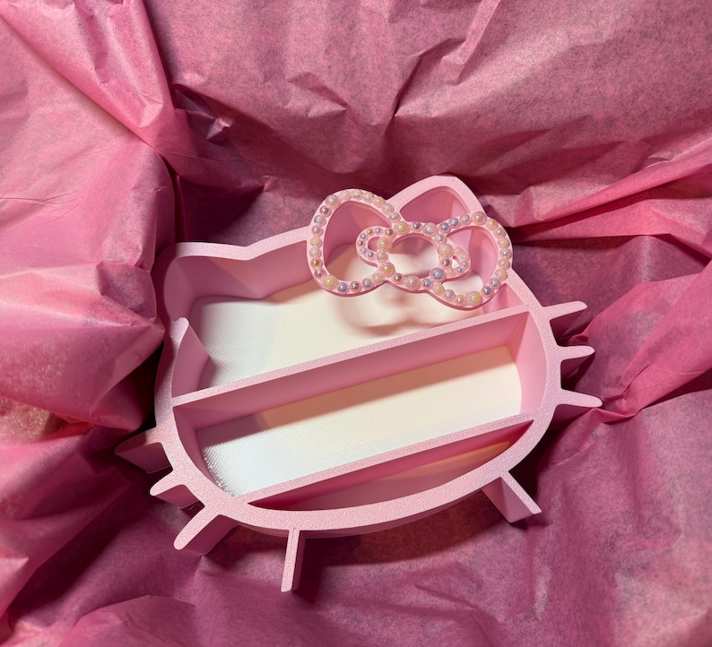 K&ouml;nnte beinhalten: Ein rosa Organizer mit drei F&auml;chern in Form eines Zeichentrickkatzenkopfes. Der Organizer hat eine schleifenf&ouml;rmige Verzierung mit kleinen, irisierenden Perlen. Er liegt auf rosa Seidenpapier.