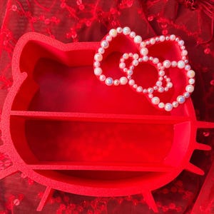 K&ouml;nnte beinhalten: Ein leuchtend rotes, Hello Kitty-f&ouml;rmiges Regal mit zwei Innenregalen. Eine perlenartige Schleife befindet sich oben. Das Regal besteht aus einem festen Material und steht auf einer roten, strukturierten Oberfl&auml;che.