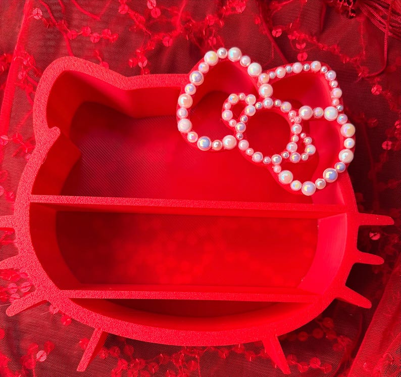 K&ouml;nnte beinhalten: Ein leuchtend rotes, Hello Kitty-f&ouml;rmiges Regal mit zwei Innenregalen. Eine perlenartige Schleife befindet sich oben. Das Regal besteht aus einem festen Material und steht auf einer roten, strukturierten Oberfl&auml;che.