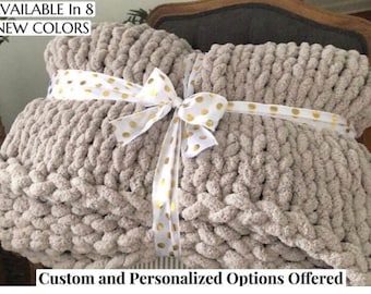 May include: A chunky knit blanket in a neutral beige color, tied with a white ribbon adorned with gold polka dots. The blanket is folded and appears soft and cozy. Text at the top says "AVAILABLE In 8 NEW COLORS".