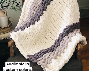 May include: A chunky knit throw blanket with a white and gray ombre pattern. The blanket is draped over a chair and has a white pom pom on the end. Available in custom colors, patterns & sizes. BAZAART