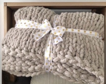 May include: A beige chunky knit throw blanket with a white ribbon tied around it. The ribbon has gold polka dots.