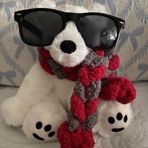 May include: A white plush polar bear wearing black sunglasses and a red, gray, and white scarf. The bear has a black nose and black eyes. The bear is sitting on a white surface.