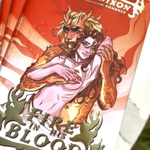 Op de afbeelding: Gestapelde boeken met een feloranje omslag. De cover toont een fantasie-illustratie van een stel, met de titel "Fire in His Blood" in goud. De naam van de auteur, Ruby Dixon, is ook zichtbaar. Thema: romantiek.