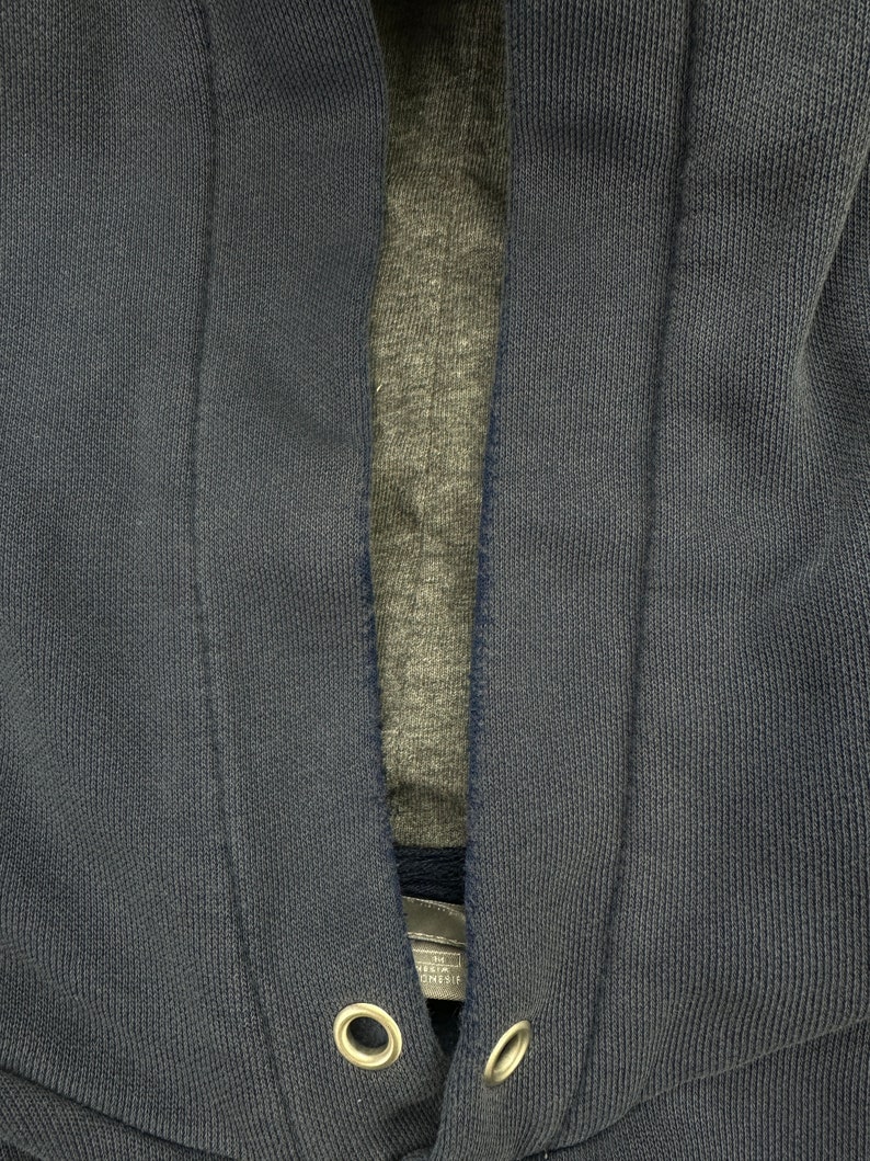 May include: Close-up of a dark blue hooded sweatshirt with a zipper closure. The sweatshirt has a gray lining and two metal eyelets near the bottom.