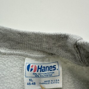 May include: A gray Hanes sweatshirt tag with white text. The tag says "XL 46-48" and "50% Cotton/Coton 50% Polyester". It also says "Made in U.S.A. Fabrique aux E-U."