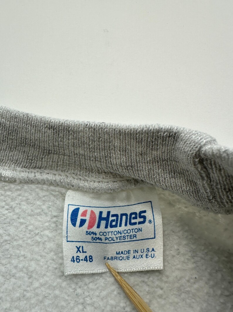 May include: A gray Hanes sweatshirt tag with white text. The tag says "XL 46-48" and "50% Cotton/Coton 50% Polyester". It also says "Made in U.S.A. Fabrique aux E-U."