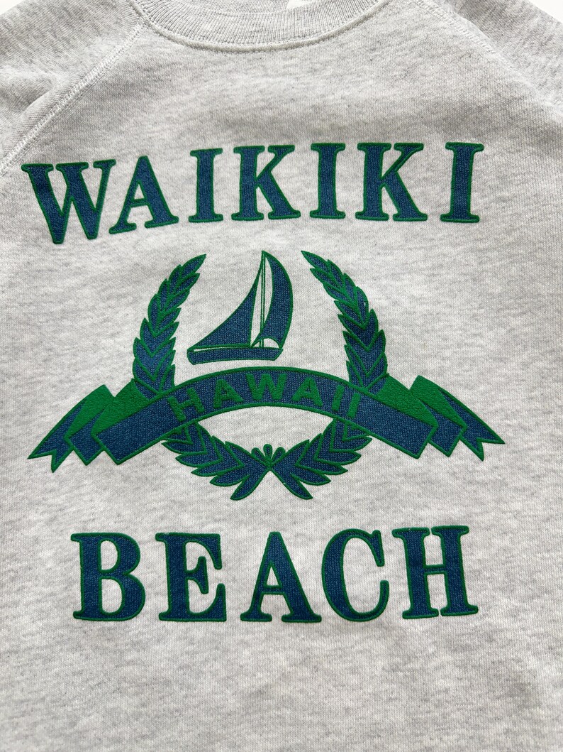 May include: A gray sweatshirt with a green and blue graphic print that says "WAIKIKI BEACH" with a sailboat and a laurel wreath.