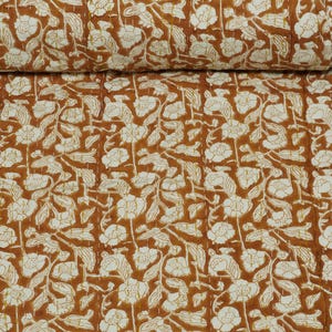 Op de afbeelding: Een opgerolde quilt met een bloemenpatroon in bruin en wit. Het uitgevouwen gedeelte toont een herhalend ontwerp van bloemen en bladeren. De rand heeft een decoratief patroon. Gemaakt van stof.