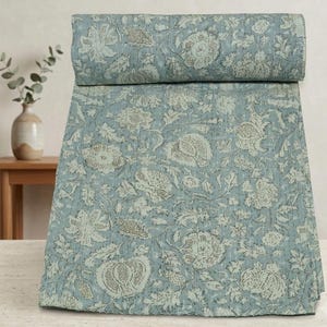 May include: A folded quilt featuring a light blue background with a repeating pattern of white and muted green floral and botanical motifs. The fabric displays visible stitching lines throughout, creating a textured appearance across the surface.