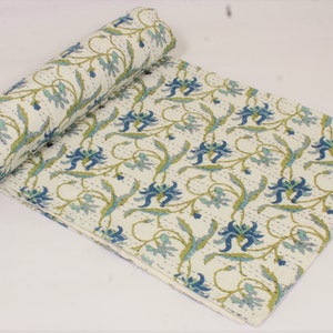 May include: A cream-colored cotton quilt featuring a repeating floral pattern in shades of blue and green. The fabric displays intricate, fine running stitch details throughout the surface, with one end of the quilt rolled into a cylinder shape.