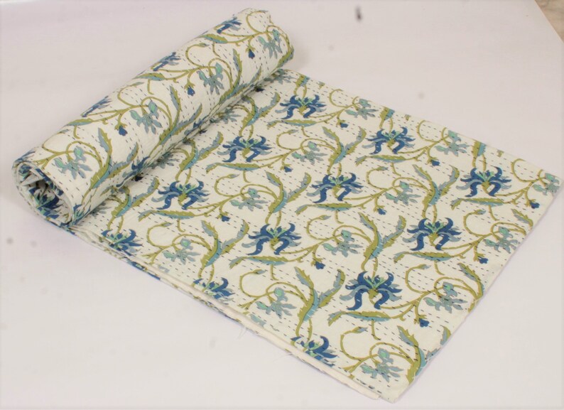 May include: A cream-colored cotton quilt featuring a repeating floral pattern in shades of blue and green. The fabric displays intricate, fine running stitch details throughout the surface, with one end of the quilt rolled into a cylinder shape.