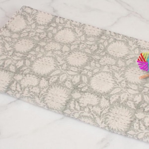 May include: A rectangular textile featuring a repeating floral pattern in light gray and off-white. The fabric displays visible running stitch details across the surface, creating a quilted texture. A small, colorful, multi-pointed star-shaped logo is attached.