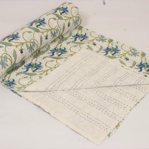 May include: A white cotton quilt featuring a repeating floral pattern in shades of blue and green. The fabric displays visible running stitch details throughout, with one end rolled up to reveal a plain white underside with parallel stitching lines.