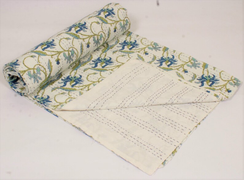 May include: A white cotton quilt featuring a repeating floral pattern in shades of blue and green. The fabric displays visible running stitch details throughout, with one end rolled up to reveal a plain white underside with parallel stitching lines.