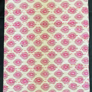 May include: A folded textile featuring a repeating pattern of pink teardrop shapes with intricate detailing on a cream background. The design is stitched with red thread, creating a textured appearance. The fabric is likely cotton or a similar material.