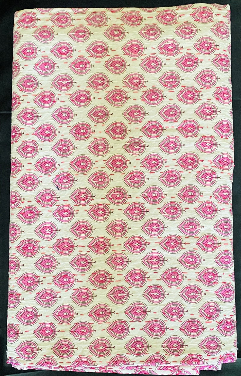 May include: A folded textile featuring a repeating pattern of pink teardrop shapes with intricate detailing on a cream background. The design is stitched with red thread, creating a textured appearance. The fabric is likely cotton or a similar material.