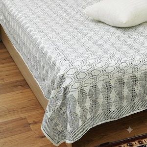 May include: A white cotton bedspread featuring a black hand-stitched kantha pattern with a central geometric motif and a border of vertical zig-zag lines. A single white pillow rests on the corner of the bedspread, which covers a wooden bed frame.
