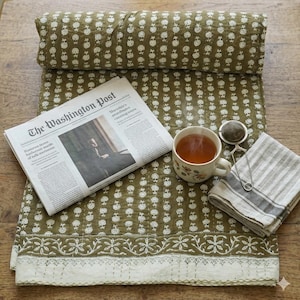 Puede incluir: Un edredón con estampado floral verde oliva con una sección enrollada en la parte superior. Un ejemplar del periódico The Washington Post está colocado sobre el edredón. También hay una taza de té floral con té, un infusor de té y una servilleta de lino a rayas.