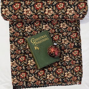 May include: A black fabric quilt featuring a repeating floral pattern in red, cream, and brown. A green book titled Christmas Traditions and a round red ornament with gold filigree details are placed on top of the folded textile.