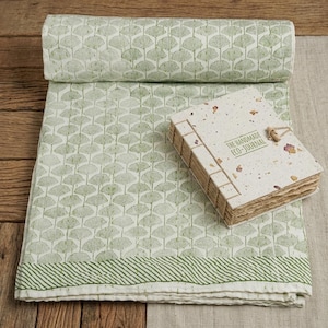 May include: A folded white cotton quilt featuring a repeating green ginkgo leaf pattern and a striped border, accompanied by a rectangular handmade eco-journal with exposed stitching and the text THE HANDMADE ECO-JOURNAL printed on the cover.
