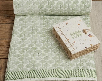 May include: A folded white cotton quilt featuring a repeating green ginkgo leaf pattern and a striped border, accompanied by a rectangular handmade eco-journal with exposed stitching and the text THE HANDMADE ECO-JOURNAL printed on the cover.