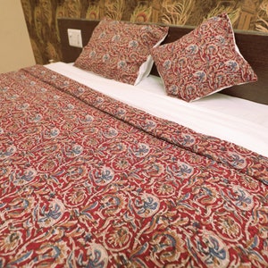 Op de afbeelding: Een bed is opgemaakt met een rood patroonquil en twee bijpassende kussenslopen. Het patroon bevat blauwe, bruine en witte bloemmotieven op een rode achtergrond. Het bed is bedekt met een wit laken.
