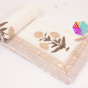 May include: Cream-colored floral quilt with brown and tan accents. The quilt features a repeating floral pattern and a decorative border. The quilt is partially rolled up, showcasing the detailed stitching and design. The text "OMRA TEXTILE" is visible.