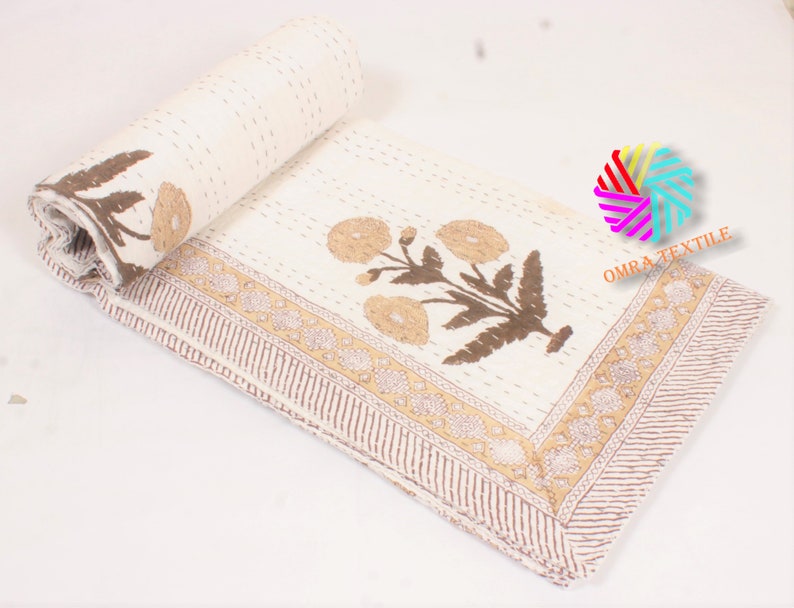 May include: Cream-colored floral quilt with brown and tan accents. The quilt features a repeating floral pattern and a decorative border. The quilt is partially rolled up, showcasing the detailed stitching and design. The text "OMRA TEXTILE" is visible.