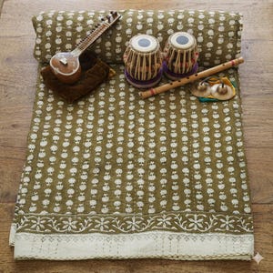 Op de afbeelding: Een olijfgroen gewatteerd textiel met een wit geblokt bloemenpatroon, gepresenteerd met een sitar, een paar tabla-trommels, een houten fluit en twee kleine koperen cimbalen die op het oppervlak van de stof rusten.