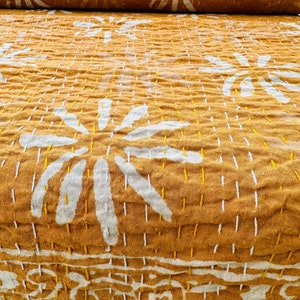 May include: Mustard-colored textile with white floral patterns and intricate stitching. The fabric is folded, revealing a rolled section. The design features large, stylized flowers and detailed border patterns.