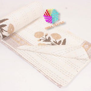 May include: Cream-colored quilt with brown floral and leaf designs. The quilt is partially rolled up, revealing the floral pattern and a decorative border. The stitching is visible throughout the quilt, adding texture. The text "OMRA TEXTILE" is visible.