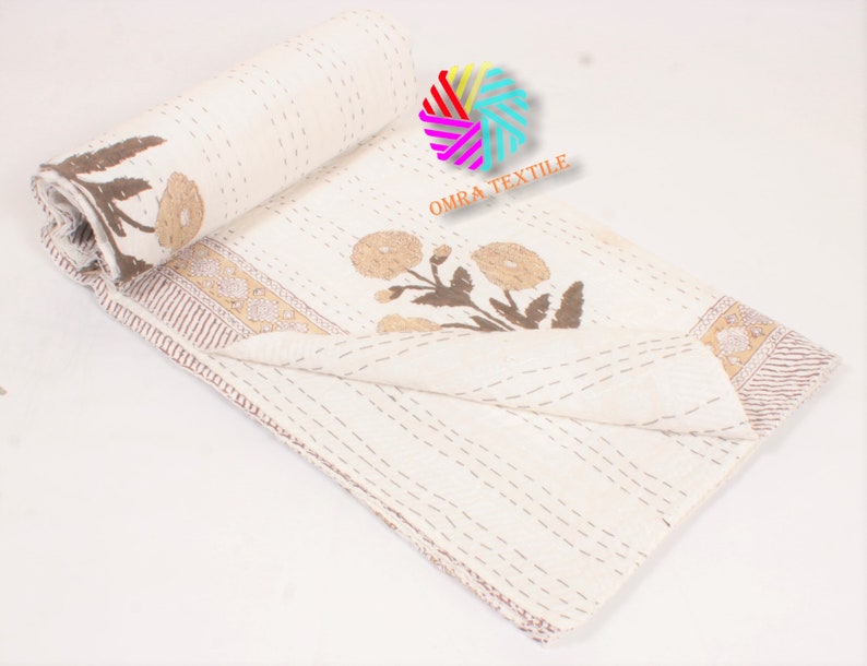 May include: Cream-colored quilt with brown floral and leaf designs. The quilt is partially rolled up, revealing the floral pattern and a decorative border. The stitching is visible throughout the quilt, adding texture. The text "OMRA TEXTILE" is visible.