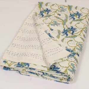 May include: A folded cotton quilt featuring a cream-colored base with a repeating floral pattern in shades of blue and green. The fabric displays visible running stitch details in dark thread, creating a textured, quilted appearance throughout.