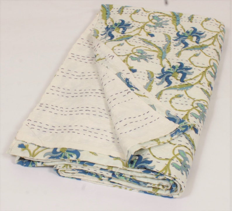 May include: A folded cotton quilt featuring a cream-colored base with a repeating floral pattern in shades of blue and green. The fabric displays visible running stitch details in dark thread, creating a textured, quilted appearance throughout.