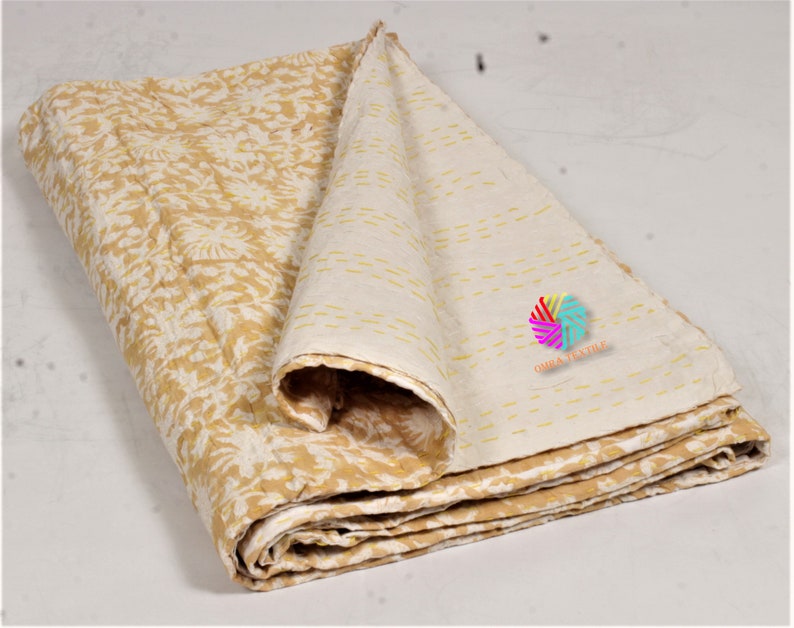 May include: A folded, reversible quilt featuring a floral design in yellow and white. The reverse side is a cream colour with yellow stitching. The quilt is made of soft fabric, suitable for a bed or sofa.
