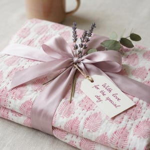 Op de afbeelding: Een cadeau verpakt in een roze en witte stof met patroon, vastgebonden met een bijpassend satijnen lint en een label met de tekst "With love for the special day". Het cadeau is versierd met lavendel en eucalyptus.