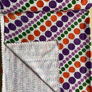 Può includere: Una trapunta bianca con un motivo ripetuto di grandi pois colorati viola, arancioni e verdi. La trapunta è piegata, rivelando un interno cucito. I bordi sono rifiniti con una cucitura decorativa.