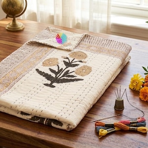 May include: A folded cream-colored quilt with a floral design and a matching pillowcase. The quilt features a brown border and a central flower motif. A small globe, flowers, and sewing supplies are arranged on a wooden table.