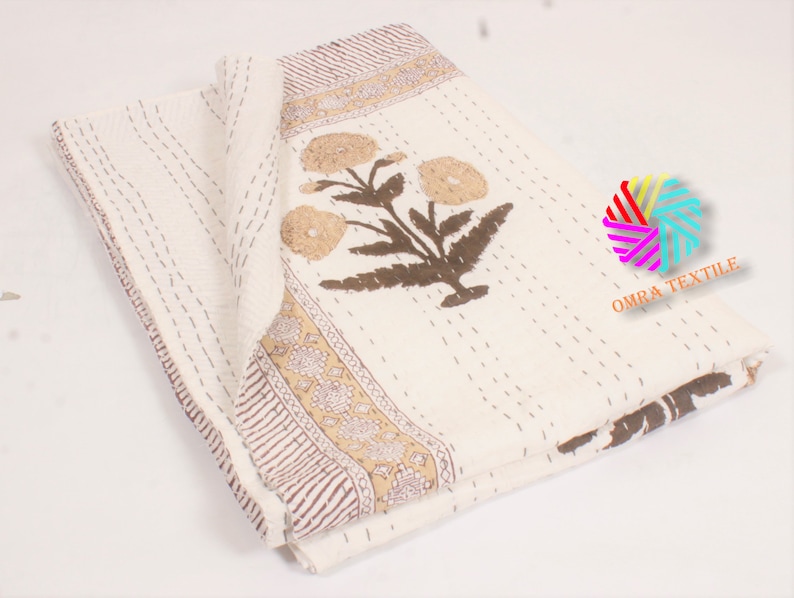 May include: Cream-colored floral quilt with brown and tan accents. The quilt features a large floral design and a decorative border. The stitching is visible throughout the quilt, adding texture and detail. The quilt is folded, showcasing its design.