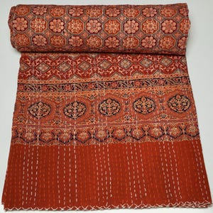 May include: A folded cotton quilt featuring a repeating geometric and floral pattern in shades of terracotta, rust, and black. The fabric displays intricate block-printed motifs and is finished with visible white running stitch kantha embroidery.