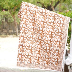 Op de afbeelding: Een wit en bruin bloemenpatroon textiel, mogelijk een deken of sprei, is over een houten staaf gedrapeerd. De stof heeft een herhalend ontwerp van gestileerde bloemen en bladeren in bruin tegen een witte achtergrond. De randen hebben een decoratieve rand.