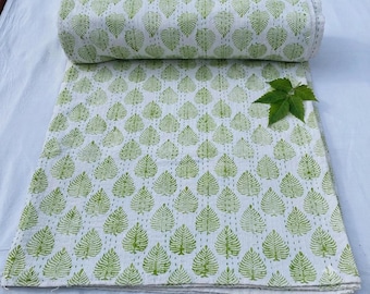May include: A white cotton quilt featuring a repeating pattern of green leaf prints and vertical running stitch lines. The fabric is folded at the top, and a single green leaf sprig is placed on the surface of the textile.