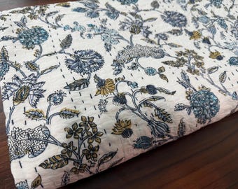 Könnte beinhalten: Gefaltetes weißes Textil mit floralem Muster in Blau-, Grün- und Gelbtönen. Das Design zeigt stilisierte Blumen und Blätter mit sichtbaren Nähten. Der Stoff wirkt weich und leicht.