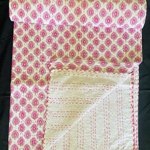May include: A rolled-up, patterned textile with a white background and a repeating pink leaf design. The unfolded portion reveals a white, quilted fabric with red stitching. The textile is likely a blanket or throw.
